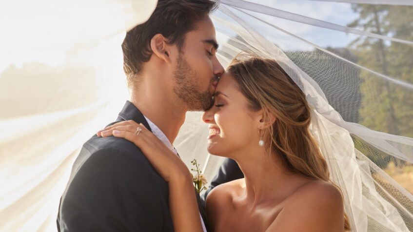 A groom kisses the bride’s forehead under her veil