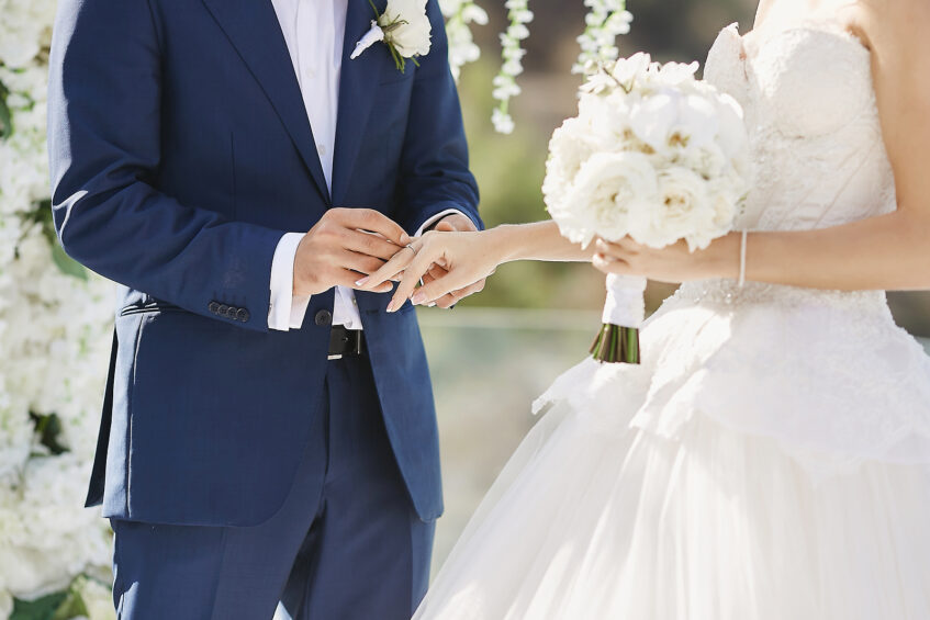 Hands with wedding rings. Modish groom putting a golden ring on the brides finger during the wedding ceremony. Loving couple, a girl in a wedding dress and handsome man in a stylish blue suit