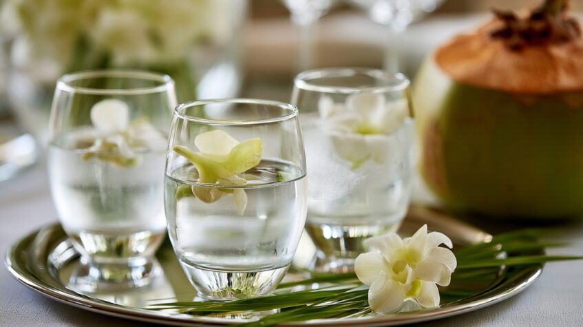 Glasses of infused water with white flowers served on a tray beside a fresh coconut.