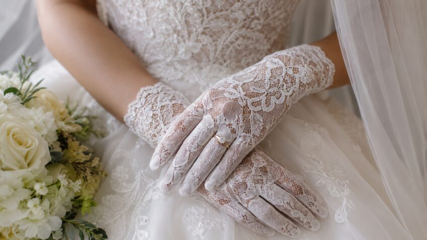 Bride’s lace gloves rest on her bridal dress beside a white bouquet