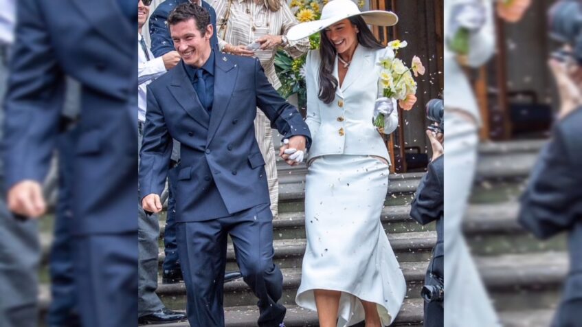 Bride in a white skirt suit and wide-brim hat walks down wedding venue steps with her partner