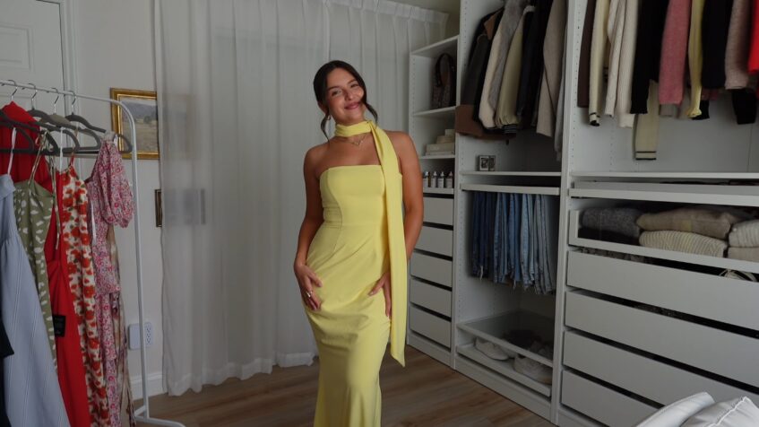 A woman wears a pale yellow scarf dress in a walk-in closet